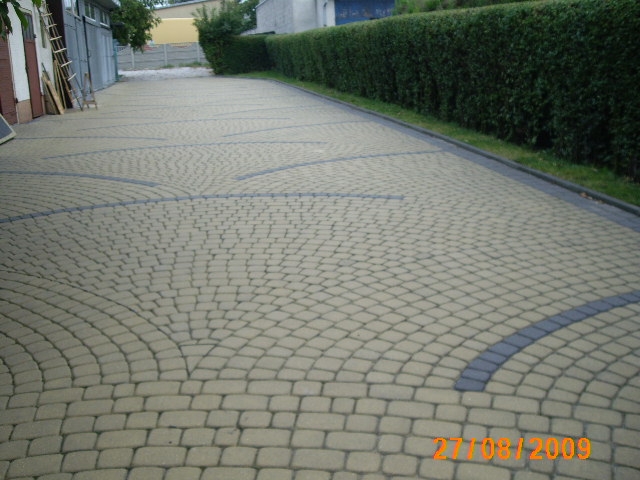 Ułożona kostka brukowa w odcieniach beżu i szarości, tworząca wzór wachlarza na długim podjeździe, obok budynku gospodarczego i żywopłotu.